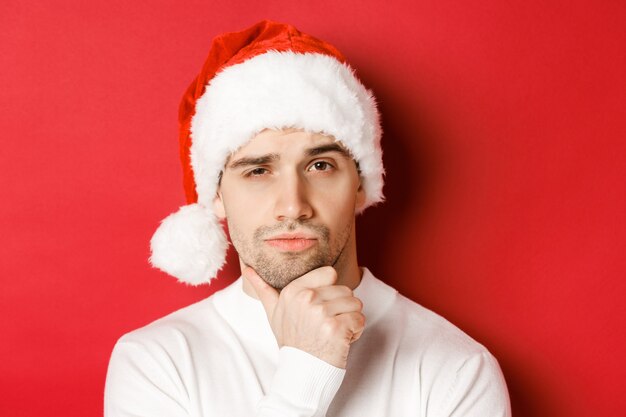 Close-up of thoughtful handsome man in santa hat, frowning and looking at camera, thinking about something, standing over red background.