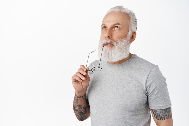 Close up of thoughtful mature bearded man biting temple of glasses looking up and thinking making choice and pondering decision standing over white background