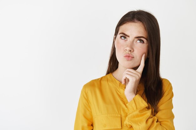 Close-up of thoughtful young woman searching solution, looking up and thinking with focused face, standing over white wall