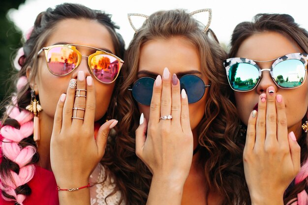 デザインされたマニキュアで創造的なサングラスをかけている3人の若い女の子のクローズアップ、2人の女性が口を覆い、1人が手で顔を隠しています。一緒にポーズをとって楽しんで幸せな友達。