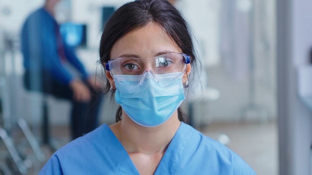 Крупным планом усталая медсестра с защитной маской от вспышки коронавируса в зале ожидания больницы. Пациент с врачом в больничной комнате осмотра.