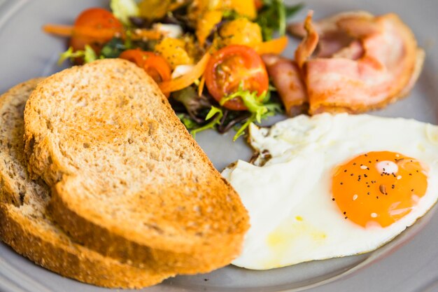 Close-up of toast; fried eggs; salad and bacon on gray ceramic plate