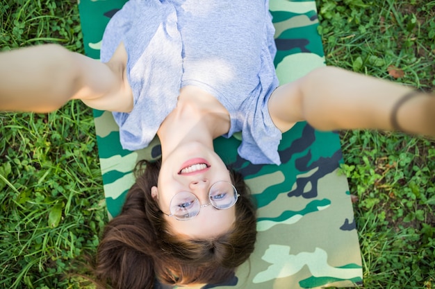 Close up top view of laughing brunette woman in eyeglasses lying on grass in park and making selfie