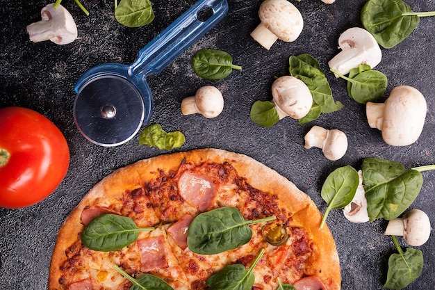 Close up top view on pizza next to mushrooms, tomatoes and leaves next to a pizza knife on dark wooden background