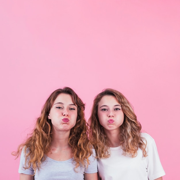 Close-up of two female friends blowing her cheeks out against pink background