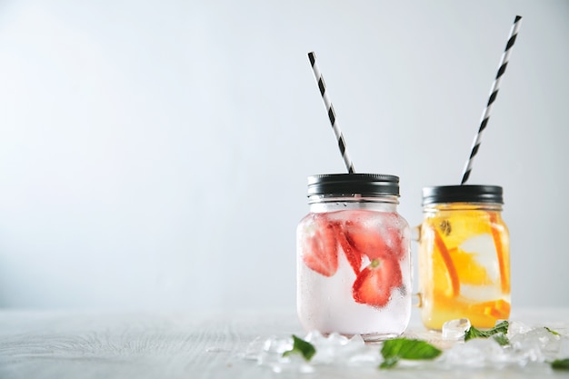 Free photo close up two fresh homemade lemonades made from sparkling water, ice, strawberry and orange. melted crashed ice and mint leaves around, striped drinking straw inside rustic jars.