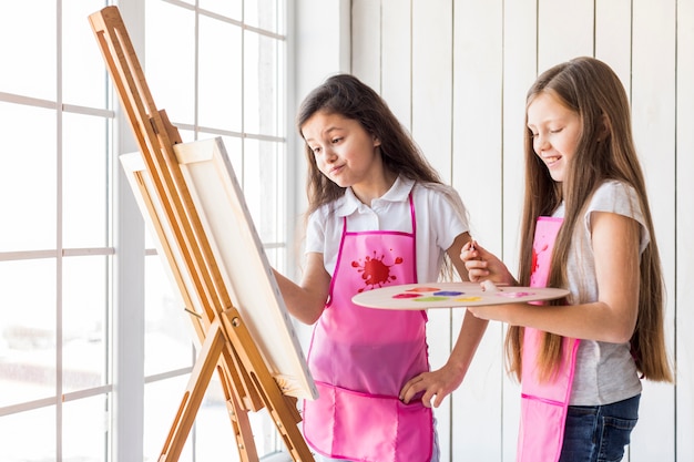 Close-up of two girls standing near the window painting on the easel with paint brush