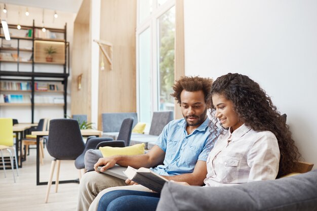 Close up of two young serious multi-ethnic students sitting on sofa in university library looking through information for exams in books, talking about university life