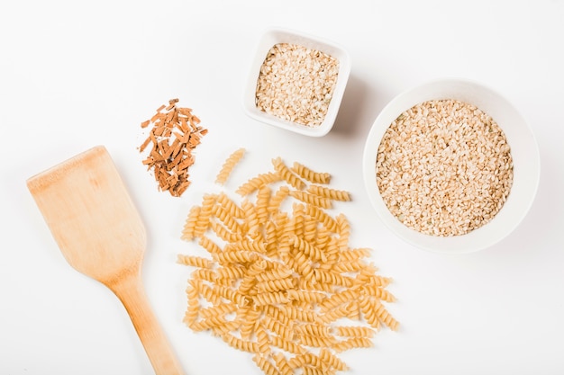 Close-up of uncooked fusilli pasta; rice and crushed cinnamon with spatula on white background