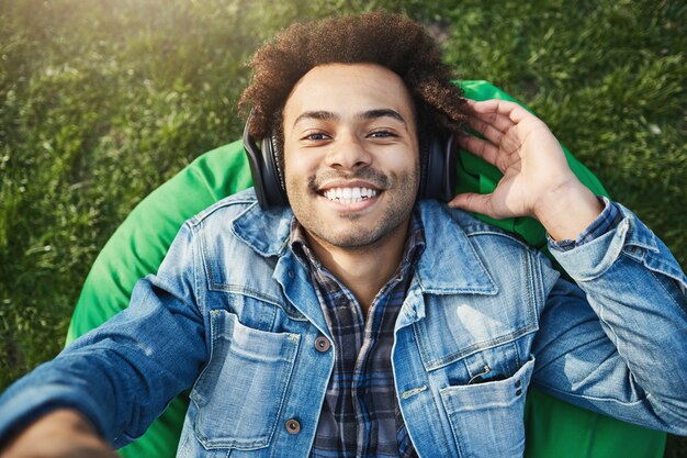 Close-up upper view shot of handsome african man with afro hairstyle extending hand towards camera while listening music via headphones, lying in park in denim coat and smiling.