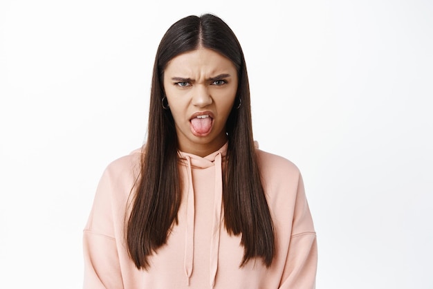 Close up of upset and moody brunette woman showing tongue and frowning grimacing from disappointment and disgusted standing against white background