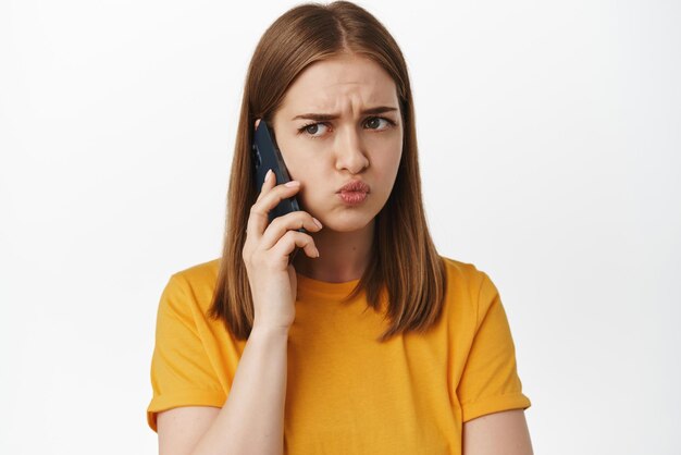 Close up of upset whining girl talking on mobile phone and grimacing disappointed with conversation hanging on hold calling someone standing over white background