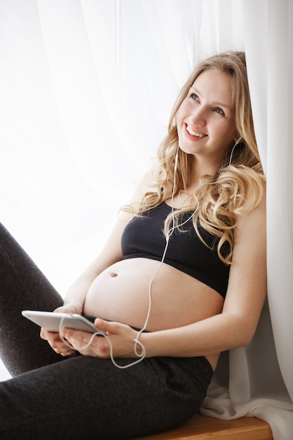 Chiuda sul ritratto verticale della madre incinta felice allegra con capelli chiari in vestiti neri che sorride, sedendosi nella camera da letto accogliente