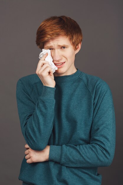 Close up vertical portrait of dramatical ginger good-looking teenager in green sweater holding napkin in hand, wipes up fake tears from face, trying to make friends feeling guilty about abusing him