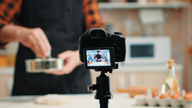 Close up of a video camera filming senior smiling man blogger in kitchen cooking. Retired blogger chef influencer using internet technology communicating on social media with digital equipment