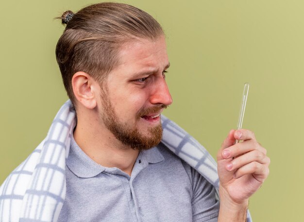 Close-up view of anxious young handsome slavic ill man wrapped in plaid holding and looking at thermometer isolated on olive green wall