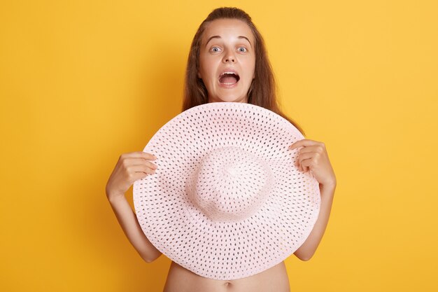 Close up view of astonished young woman with widely opened mouth covering her breasts with straw hat