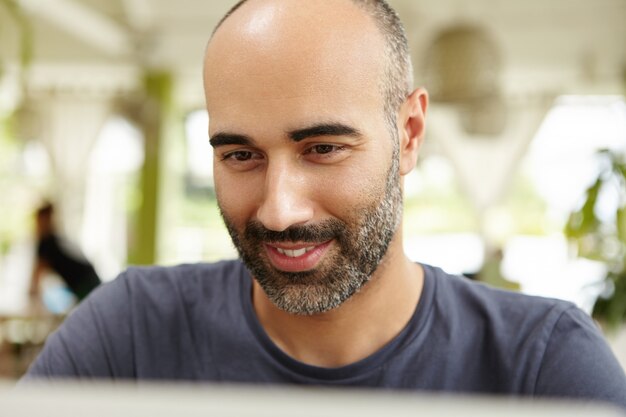 Close up view of attractive adult male with beard sitting at open terrace, typing on laptop, looking at screen with interested smile, using wi-fi to communicate online while away on vacations