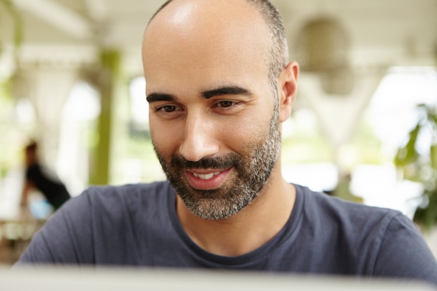 ひげがオープンテラスに座っている、ラップトップで入力する、興味のある笑顔で画面を見ている、休暇中にオンラインで通信するためにwi-fiを使用している魅力的な成人男性のクローズアップ表示