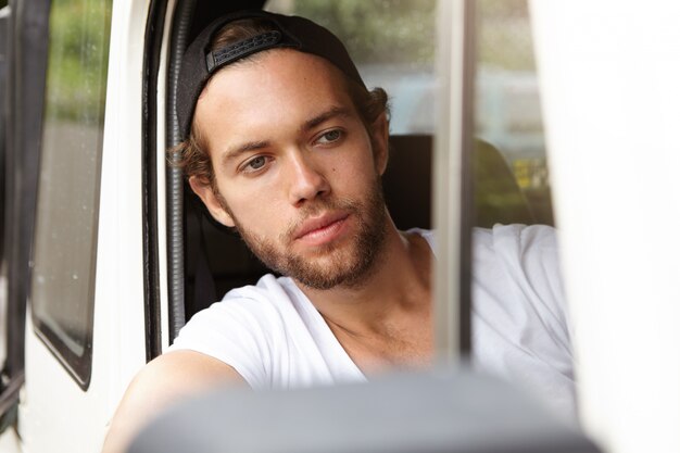 Close up view of attractive young bearded man in snapback sitting inside cabin of his white jeep