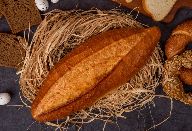 Close-up view of baguette on straw surface with different breads and eggs on maroon background