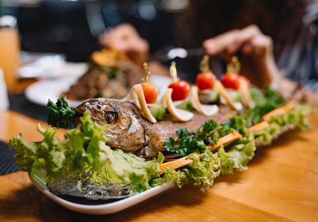 Close up view of baked seabass fish decorated with cherry tomatoes and lemon slices on lettuce on a wooden platter