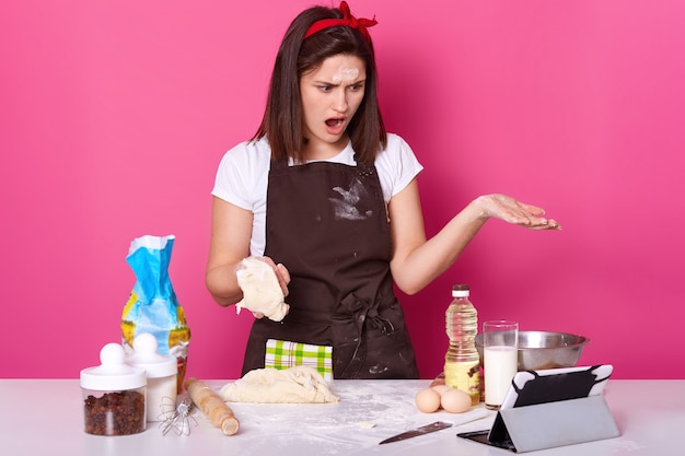 Close up view of baker kneading dough for homemade bread, has astonised facial expression, stands with open mouth, uses ipod for serching better recipe in Internet. Copy space for your advertisment.