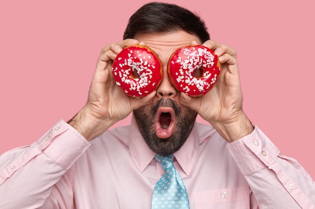 Крупным планом вид бородатого мужчины держит красные пончики на глазах, держит широко открытым рот