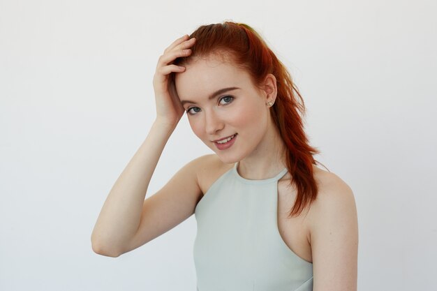 Close up view of beautiful Caucasian teenage woman smiling joyfully tying her long red hair in ponytail. Young Caucasian female model