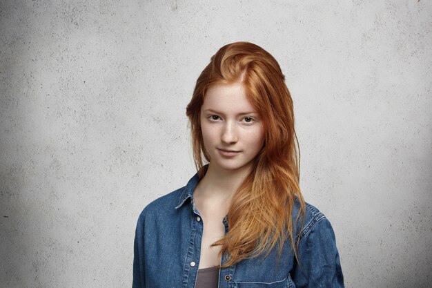 Close up view of beautiful young Caucasian woman with ginger hair and freckles wearing stylish clothes looking  with faint smile, posing against gray wall.