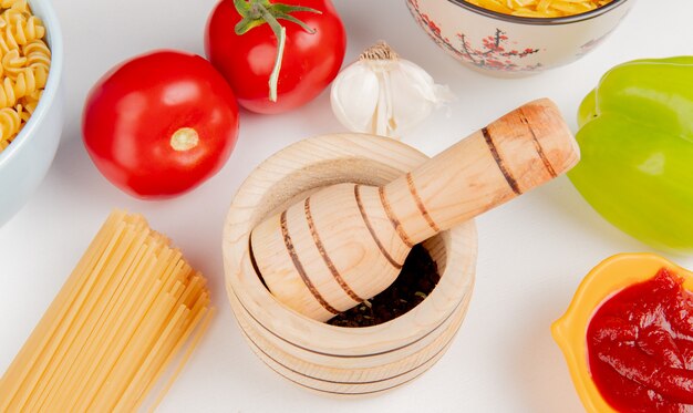 Close-up view of black pepper seeds in garlic crusher with macaronis as rotini and vermicelli tomatoes ketchup garlic pepper on white table