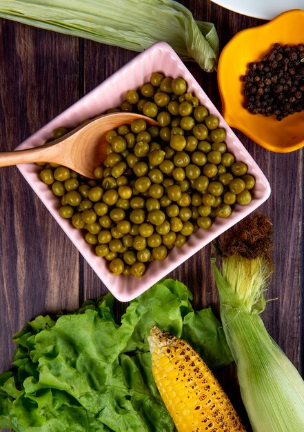 Close-up view of bowl of green peas with lettuce corns and black pepper on wooden surface