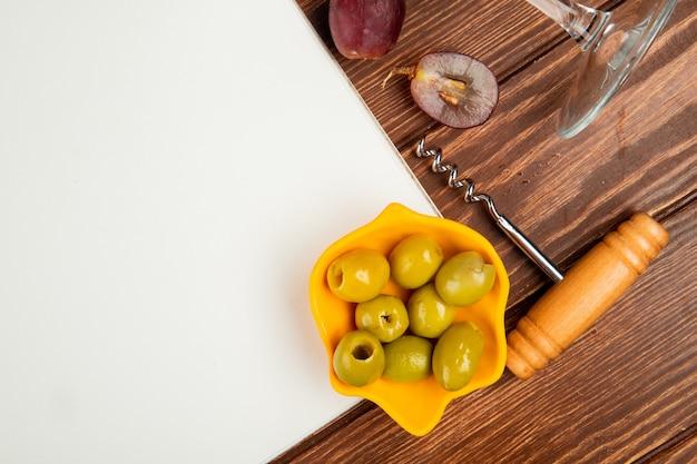 close-up view of bowl of olive and note pad with grape corkscrew on wooden background with copy space