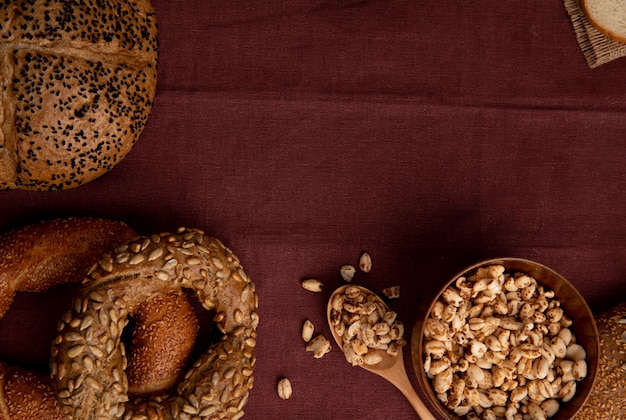 Close-up view of breads as seeded cob bagel with bowl of corns and spoonful of corns on burgundy background with copy space