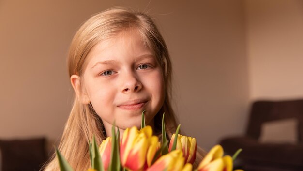 Close up view of charming young girl sitting at home with flowers and smile at camera