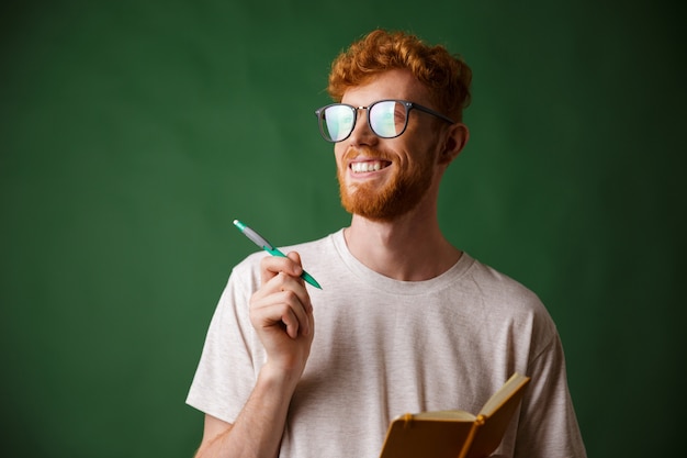 Close-up view of cheerful bearded young man in white tshirt holding a notebook and a pen