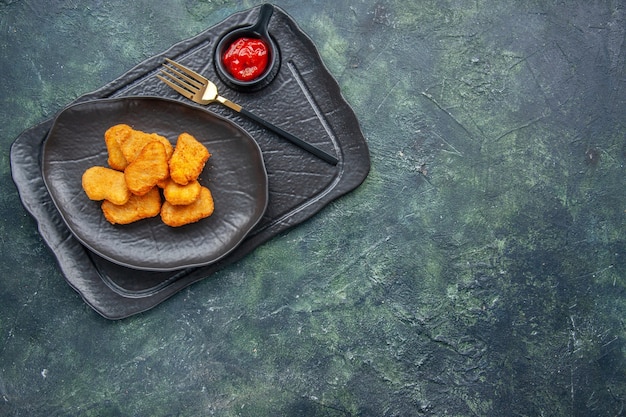Vista ravvicinata delle pepite di pollo su un piatto nero e ketchup a forchetta sul lato destro su vassoio di colore scuro