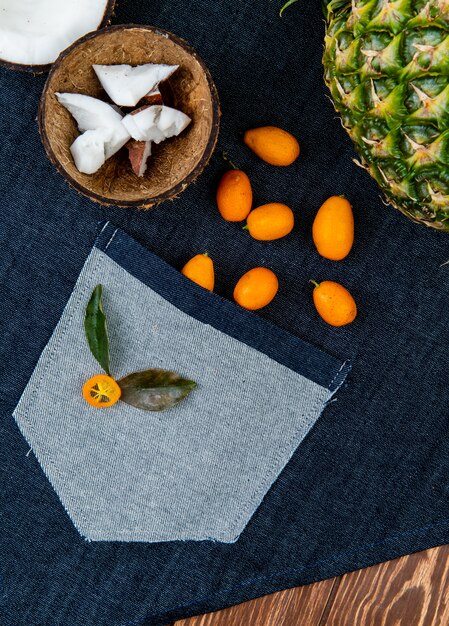 Close-up view of citrus fruits as half cut coconut with coconut slices in shell kumquats pineapple with leaves on jeans cloth and wooden background