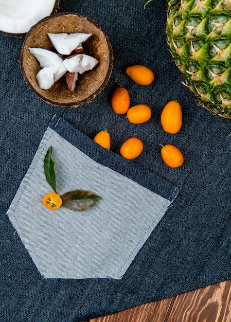 Close-up view of citrus fruits as half cut coconut with coconut slices in shell kumquats pineapple with leaves on jeans cloth and wooden background