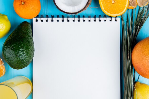 Close-up view of citrus fruits as tangerine avocado coconut lemon and lemon juice with note pad on center on blue background with copy space