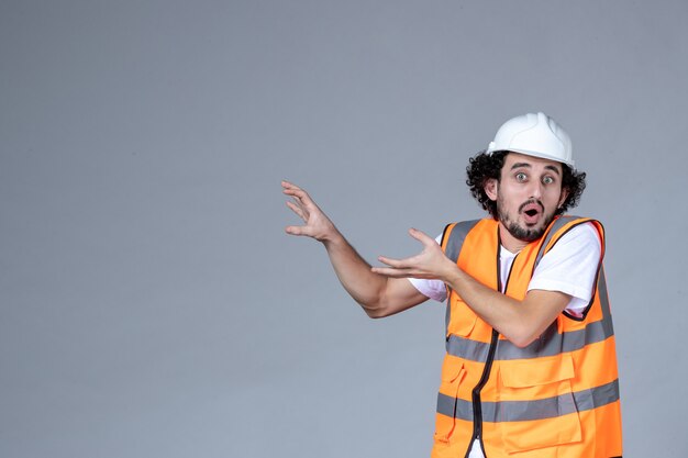 Close up view of concerned male architect in warning vest with safety helmet and pointing something on the right side on gray wave wall