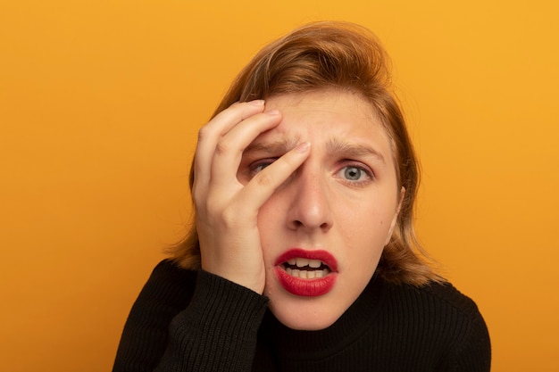 Close-up view of concerned young blonde woman looking at front putting hand on head isolated on orange wall