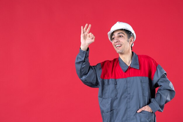 Close up view of confident young architect in uniform with hard hat and making perfect gesture on isolated red wall