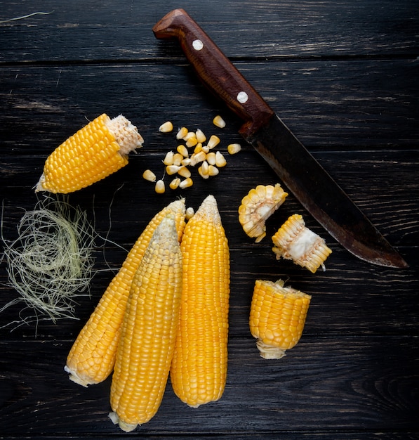 Close-up view of cooked whole and cut corns corn seeds with silk and knife on black surface
