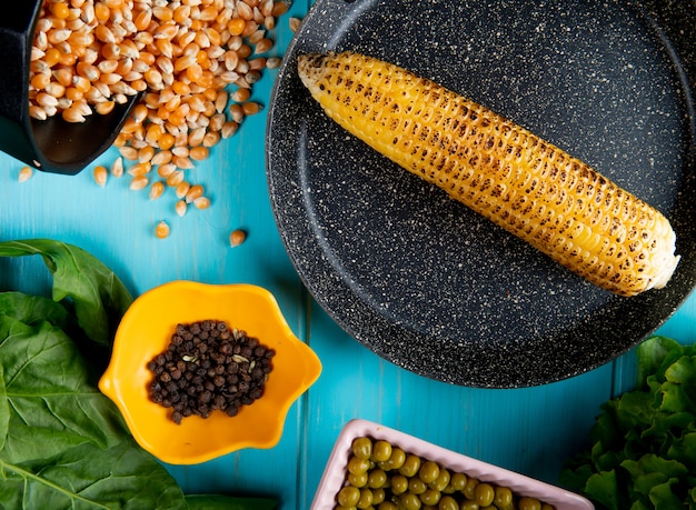 Close-up view of corn cob in pan with corn seeds black pepper seeds and lettuce on blue surface