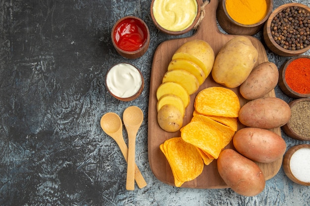 Close up view of crispy chips and uncooked potatoes on wooden cutting board and different spices on gray table