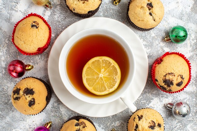 Close up view of a cup of black tea with lemon among freshly baked delicious small cupcakes and decoration accessories on ice surface