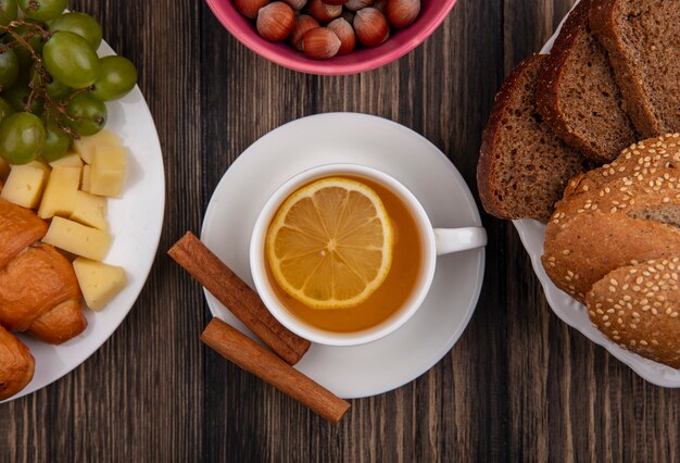 Close-up view of cup of hot toddy with cinnamon on saucer and nuts with cheese grape croissant and plate of bread slices on wooden background