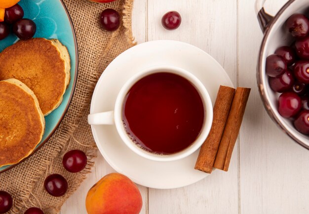 ソーサーにお茶とシナモン、皿にさくらんぼ、荒布にアプリコットさくらんぼ、木製の背景にさくらんぼのボウルのパンケーキの拡大図