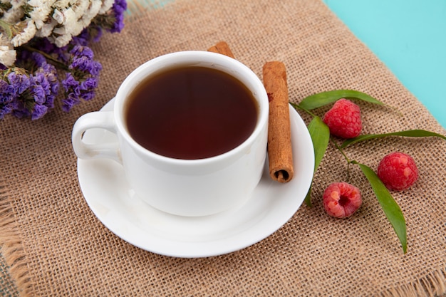 Крупным планом вид чашки чая и корицы на блюдце с малиной и листьями и цветами на вретище